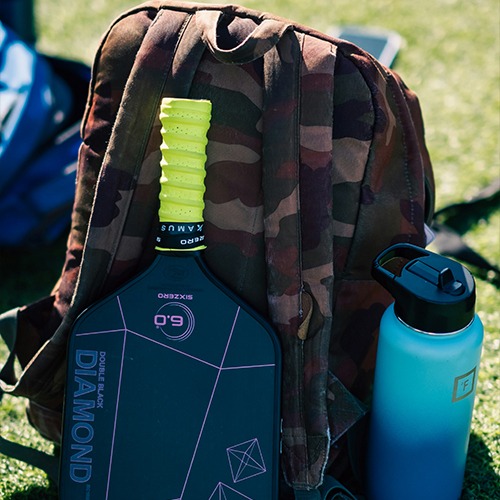 Diamond pickleball paddle rests against a camouflage backpack, with a water bottle nearby. This setup suggests readiness for outdoor sports.