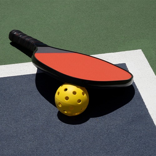 A bright red pickleball paddle rests on a blue court with a yellow perforated ball, indicating readiness for a game of pickleball.
