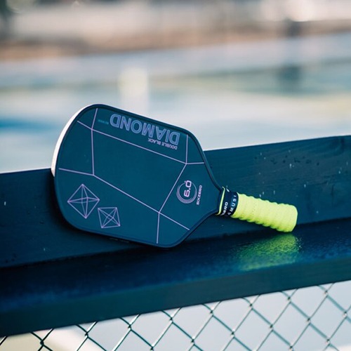 Pickleball paddle resting on a fence, featuring a sleek black design with vibrant yellow grip, highlighting its modern style and functionality.