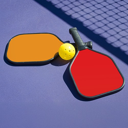 Two colorful pickleball paddles in orange and red, accompanied by a yellow pickleball, resting on a purple court surface with a net in the background.
